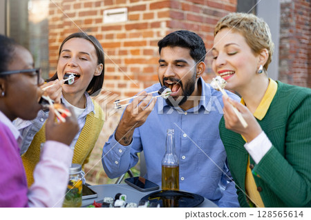 A diverse group of friends enjoys a sushi meal together, savoring the flavors and company outdoors. A diverse group of friends enjoys a sushi meal together, savoring the flavors and company outdoors. 128565614