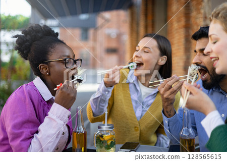 A diverse group of friends enjoys sushi outdoors, a moment of shared joy and culinary delight. A diverse group of friends enjoys sushi outdoors, a moment of shared joy and culinary delight. 128565615