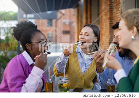 Diverse group of friends enjoying sushi outdoors, laughing and having a good time during a casual lunch. Diverse group of friends enjoying sushi outdoors, laughing and having a good time during a casual lunch. 128565616