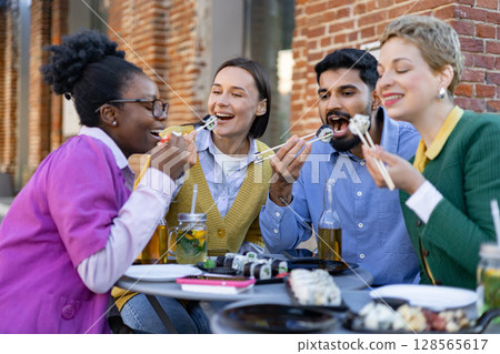 A diverse group of friends enjoys sushi and drinks together at an outdoor restaurant, laughing and having fun. A diverse group of friends enjoys sushi and drinks together at an outdoor restaurant, laughing and having fun. 128565617