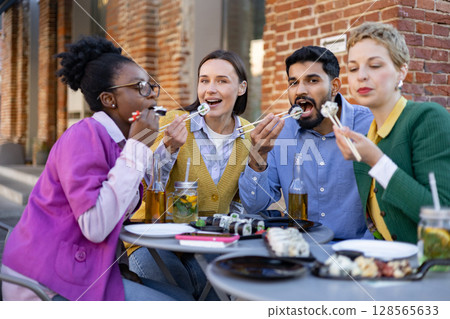 A diverse group of friends enjoys a meal of sushi together outdoors, laughing and eating. A diverse group of friends enjoys a meal of sushi together outdoors, laughing and eating. 128565633