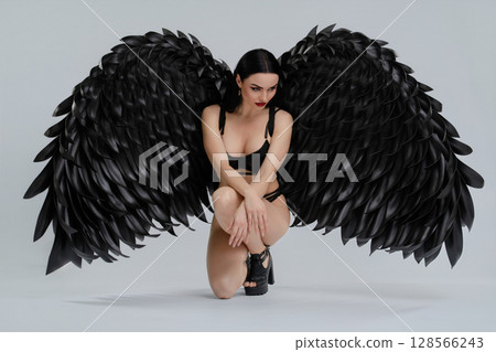 Elegant woman in black lingerie poses with dramatic wings in a studio setting Elegant woman in black lingerie poses with dramatic wings in a studio setting 128566243