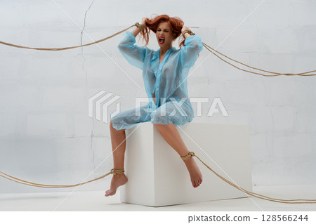 Red-haired woman tied with rope expresses emotions in a minimalistic setting Red-haired woman tied with rope expresses emotions in a minimalistic setting 128566244