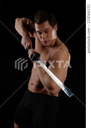 Dynamic pose of a muscular man wielding a sword in artistic lighting setup Dynamic pose of a muscular man wielding a sword in artistic lighting setup 128566331