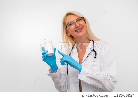 Middle aged blonde therapist in white coat, gloves, and stethoscope holding a bottle of vitamins. Isolated on white background with copy space Middle aged blonde therapist in white coat, gloves, and stethoscope holding a bottle of vitamins. Isolated on white background with copy space 128566824