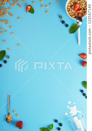Scattered cereal rings, strawberries, blueberries, mint leaves, spoons, and spilled milk blue background. Copyspace 128567140