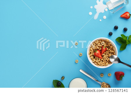 Banner with cereal rings, strawberries, blueberries, mint leaves, spoons, and spilled milk on blue background. Copyspace 128567141