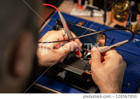 Technician repairing smartphone battery using soldering tools in a tech workshop during daylight hours 128567950