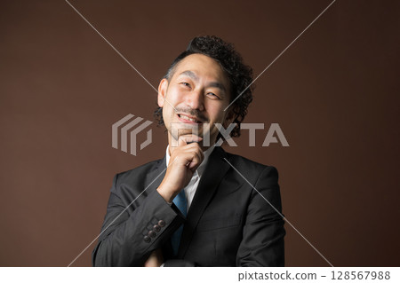 一位商人穿著西裝，手托著下巴，在棕色背景下思考 128567988