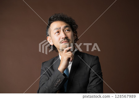 一位商人穿著西裝，手托著下巴，在棕色背景下思考 128567989