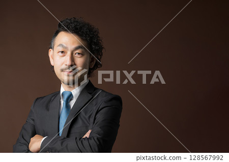 一位穿著西裝、微笑的商人，雙臂交叉，背景為棕色，象徵著信任和可信度，他正看著鏡頭 128567992
