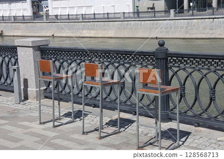 A row of chairs with wooden seats covered with snow near a metal fence on the canal embankment. 128568713