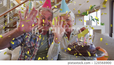 Happy caucasian senior couple in party hats making birthday image call with cake and gold confetti 128568900