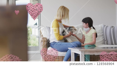 Image of hearts over caucasian woman and her daughter wearing face masks playing with pet dog 128568904