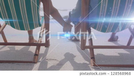 Image of light moving over senior couple on beach deckchairs holding hands 128568968
