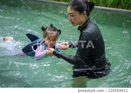 mother teaching toddler girl swimming in pool and wearing life jacket 128569421