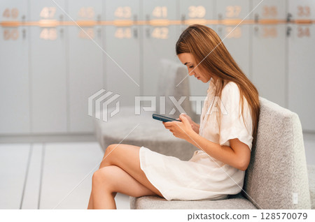 Sitting with smartphone in hands. Woman is in the gym changing room with lockers 128570079