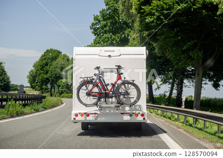 Rear view of a camper van driving on scenic road with two electric bicycles mounted on back rack 128570094