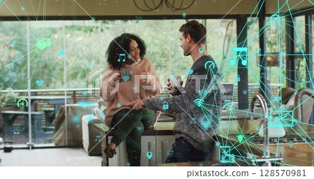 Image of network of media icons over happy biracial couple having coffee talking at home 128570981