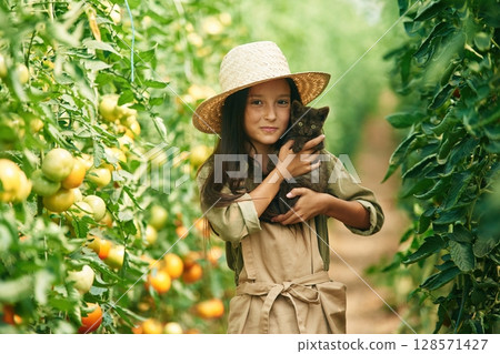 Holding cute kitten in hands. Little girl is in the garden with tomatoes 128571427