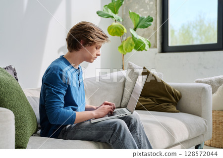 Handsome serious teenage boy sitting at home on the couch typing on laptop 128572044