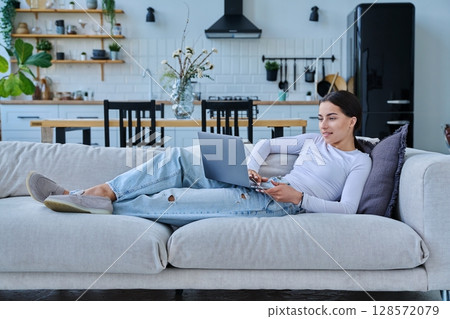 Portrait of young woman lying on sofa typing on laptop, in home interior Portrait of young woman lying on sofa typing on laptop, in home interior 128572079