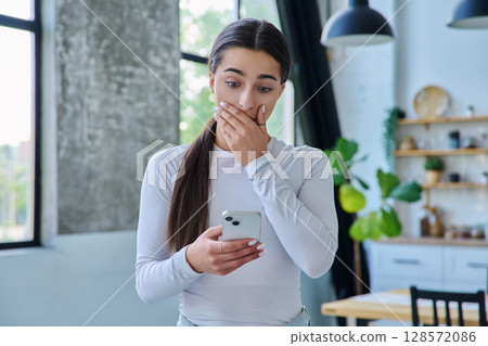 Young sad stressed woman standing at home using smartphone Young sad stressed woman standing at home using smartphone 128572086