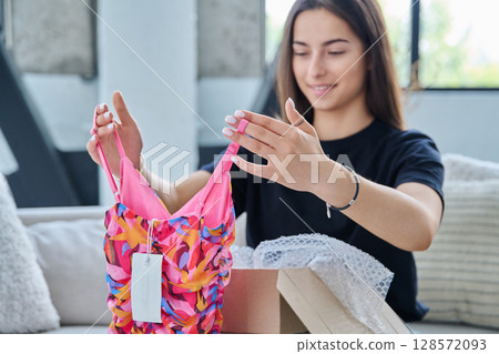 Young woman unpacking cardboard box parcel with online order, looking at new clothes 128572093