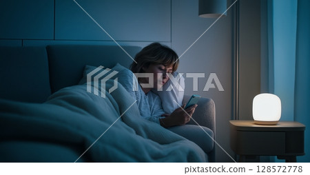 Young Woman Using Smartphone While Resting on a Couch Under a Blanket in a Softly Lit Room at Night 128572778