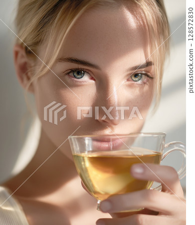 Close-up portrait of a woman with striking eyes, enjoying a warm cup of tea bathed in natural sunlight, evoking feelings of calm, wellness, and a tranquil start to the day Close-up portrait of a woman with striking eyes, enjoying a warm cup of tea bathed in natural sunlight, evoking feelings of calm, wellness, and a tranquil start to the day 128572830