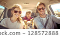 A joyful family, including parents, a child, and a small dog, all wearing sunglasses and smiling broadly from inside a sunlit car, capturing a happy road trip moment 128572888