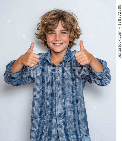 Energetic young boy gives a double thumbs-up, showcasing a positive, approving gesture. His blue eyes, genuine smile convey enthusiasm, success, perfect for conveying approval, achievement Energetic young boy gives a double thumbs-up, showcasing a positive, approving gesture. His blue eyes, genuine smile convey enthusiasm, success, perfect for conveying approval, achievement 128572889