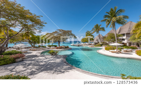 Expansive, pristine lagoon-style swimming pool. White sandy beach with lush tropical foliage, palm trees, with overwater bungalows, ocean distance. Concept of luxury, tranquility, vacation paradise 128573436