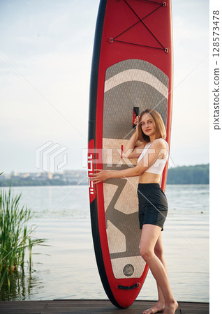Woman is standing with sup board against the lake Woman is standing with sup board against the lake 128573478