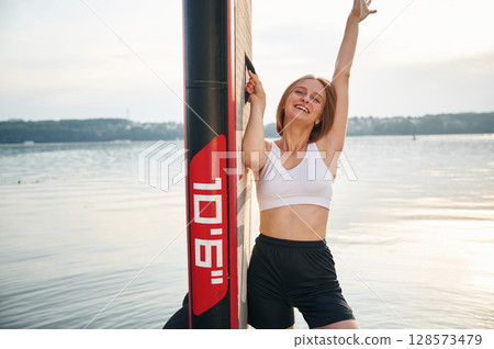 Smiling and enjoying the summer. Woman is standing with sup board against the lake Smiling and enjoying the summer. Woman is standing with sup board against the lake 128573479