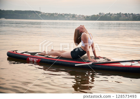 Doing fitness exercises. Young woman is on sup boards in the lake Doing fitness exercises. Young woman is on sup boards in the lake 128573493
