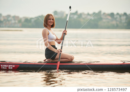 Forest and houses on background. Young woman is on sup boards in the lake Forest and houses on background. Young woman is on sup boards in the lake 128573497