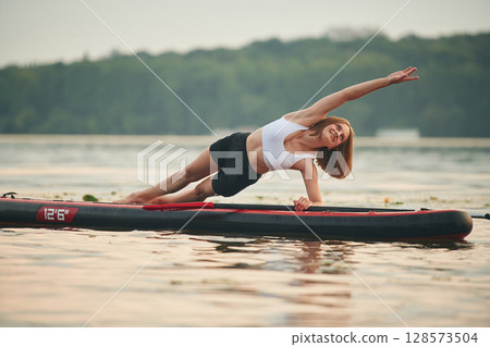 Doing fitness exercises. Young woman is on sup boards in the lake Doing fitness exercises. Young woman is on sup boards in the lake 128573504
