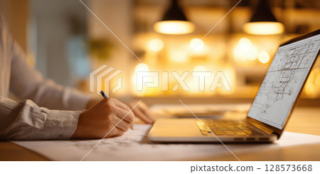 Focused individual's hands are shown meticulously drawing on blueprints, with laptop displaying architectural plans open beside them. Concept of professional work in architecture, design, engineering 128573668