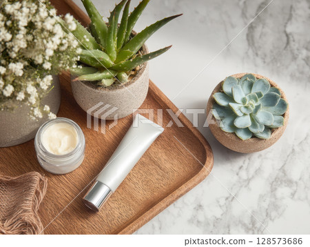 Natural skincare products and plants on a wooden tray set on a marble surface. It features a jar of cream, a white tube, a small pot with white flowers, and two potted succulents 128573686