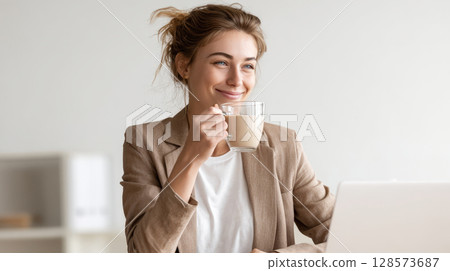 smiling woman with her hair in a bun, wearing a beige blazer over a white top, holds a clear glass mug of coffee. She is looking away thoughtfully with a pleasant expression, with a laptop 128573687