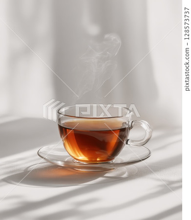 Translucent glass cup filled with amber-colored herbal tea, with gentle steam rising, placed on matching glass saucer, bathed in natural sunlight creating elegant shadows on textured white background 128573737