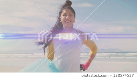 Image of lights over smiling hispanic woman picking up rubbish from beach 128574537