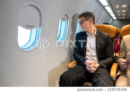 Businessman in a dark blazer and glasses thoughtfully out of airplane window while seated in cabin 128576306