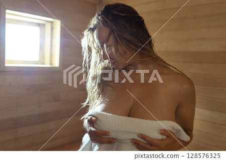 A young woman relaxes and sweats in a hot sauna wrapped in a towel. girl in the sauna. Finnish sauna interior, classic wooden sauna with hot steam. Russian bath. 128576325