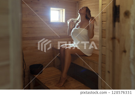 A young woman relaxes and sweats in a hot sauna wrapped in a towel. girl in the sauna. Finnish sauna interior, classic wooden sauna with hot steam. Russian bath. A young woman relaxes and sweats in a hot sauna wrapped in a towel. girl in the sauna. Finnish sauna interior, classic wooden sauna with hot steam. Russian bath. 128576331