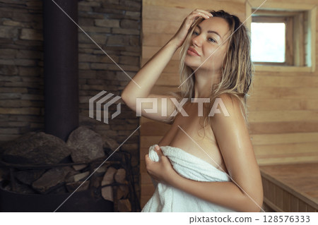 A young woman relaxes and sweats in a hot sauna wrapped in a towel. girl in the sauna. Finnish sauna interior, classic wooden sauna with hot steam. Russian bath. A young woman relaxes and sweats in a hot sauna wrapped in a towel. girl in the sauna. Finnish sauna interior, classic wooden sauna with hot steam. Russian bath. 128576333