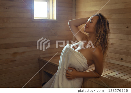 A young woman relaxes and sweats in a hot sauna wrapped in a towel. girl in the sauna. Finnish sauna interior, classic wooden sauna with hot steam. Russian bath. 128576336