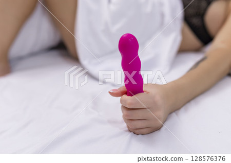 Woman in bed under sheets holding vibrator in hand 128576376