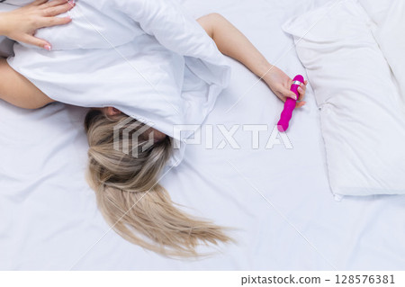 Woman in bed under sheets holding vibrator in hand 128576381